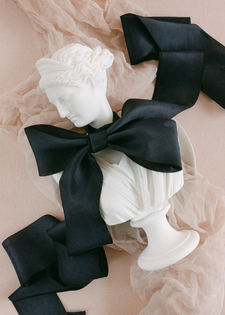 Large black silk ribbon bow tied around a Greco-Roman inspired bust, placed on soft neutral textiles
