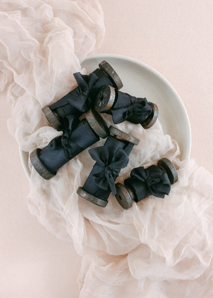 Hand-dyed black silk ribbon wrapped around vintage wooden spools, styled on soft ivory fabric for an elegant flatlay