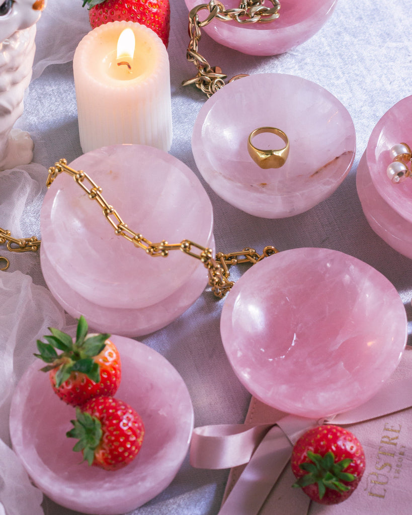 Pink gemstone rose quartz bowls with gold jewelry and strawberries on a textured surface