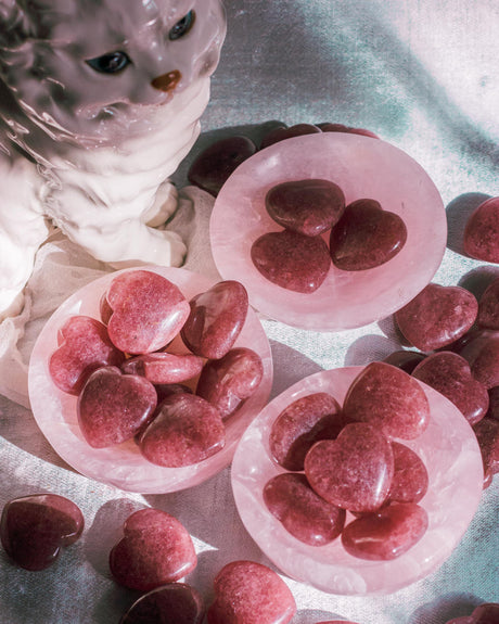 Hand-polished Strawberry Quartz heart palm stones in crystal dish, photographed on a blue surface, showcasing its smooth texture and rosy hues.