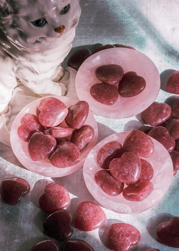 Hand-polished Strawberry Quartz heart palm stones in crystal dish, photographed on a blue surface, showcasing its smooth texture and rosy hues.