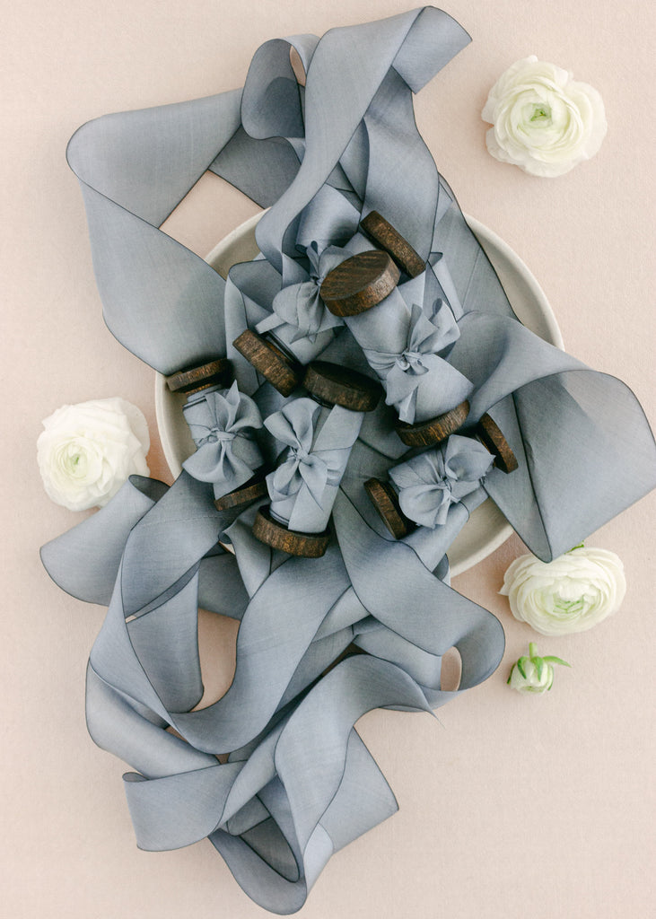Romantic styling of dusty blue silk ribbon wrapped around vintage spools, displayed with white flowers on a soft backdrop