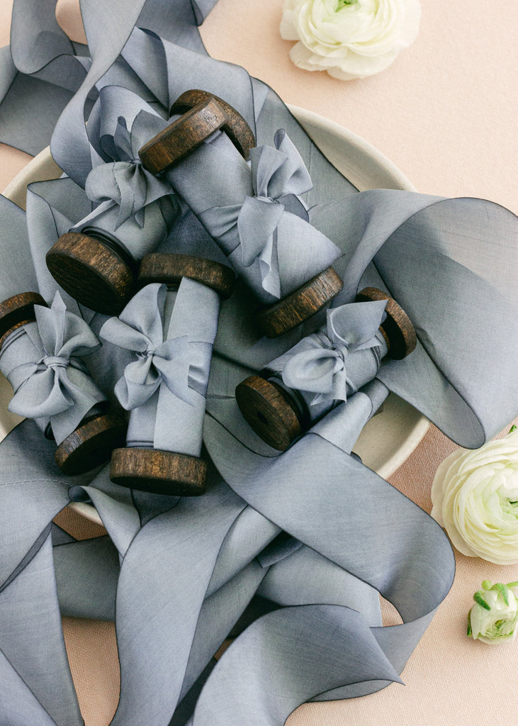 Romantic styling of dusty blue silk ribbon wrapped around vintage spools, displayed with white flowers on a soft backdrop