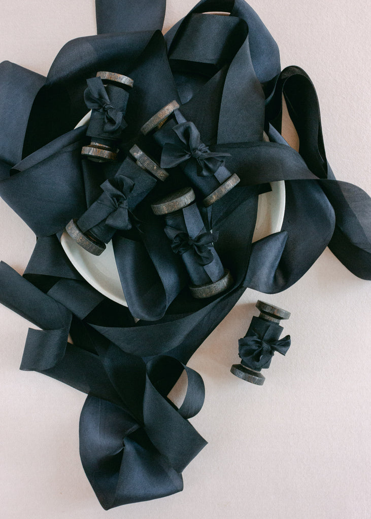 Luxury hand-dyed black silk ribbon wrapped around vintage wooden spools, styled on ceramic plate on fabric background