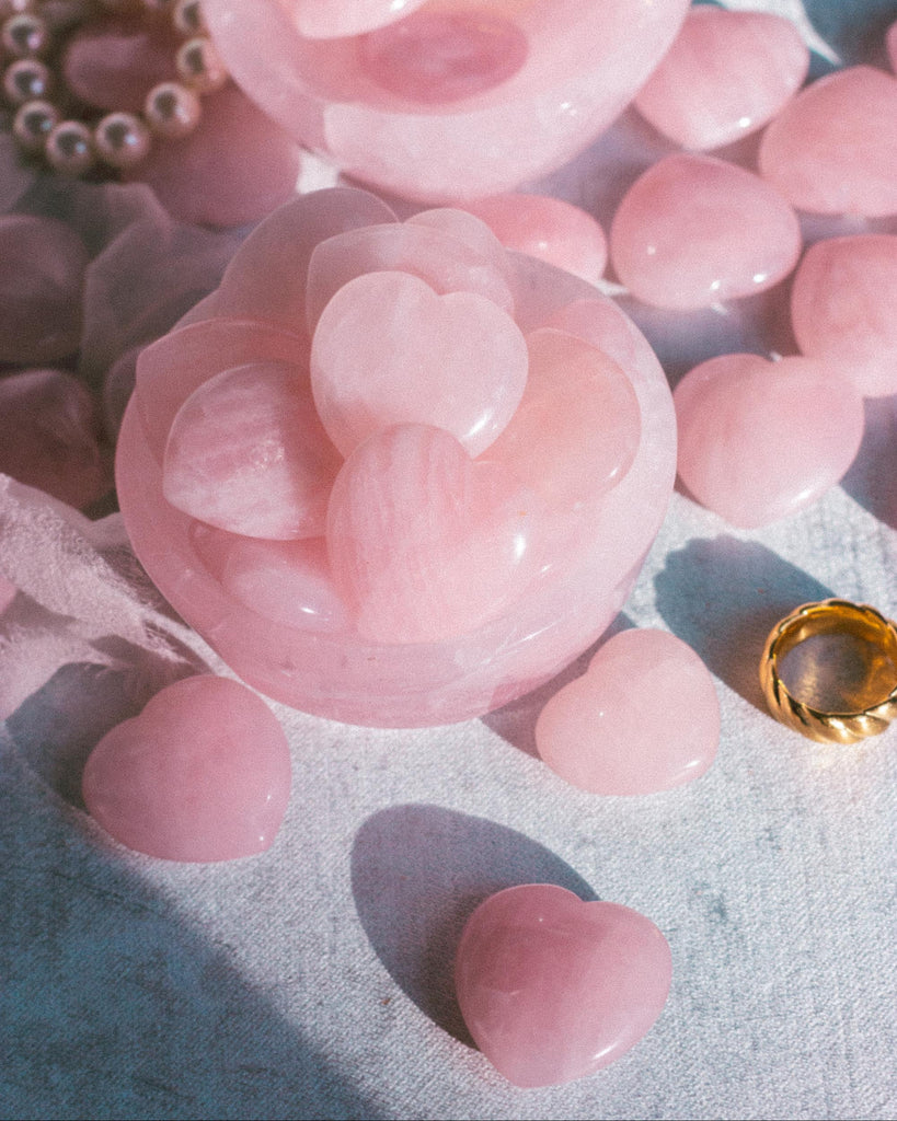 Polished Rose Quartz crystal hearts arranged inside a delicate rose quartz bowl, with their soft pink hues glowing against a rich light blue velvet background and gold ring.