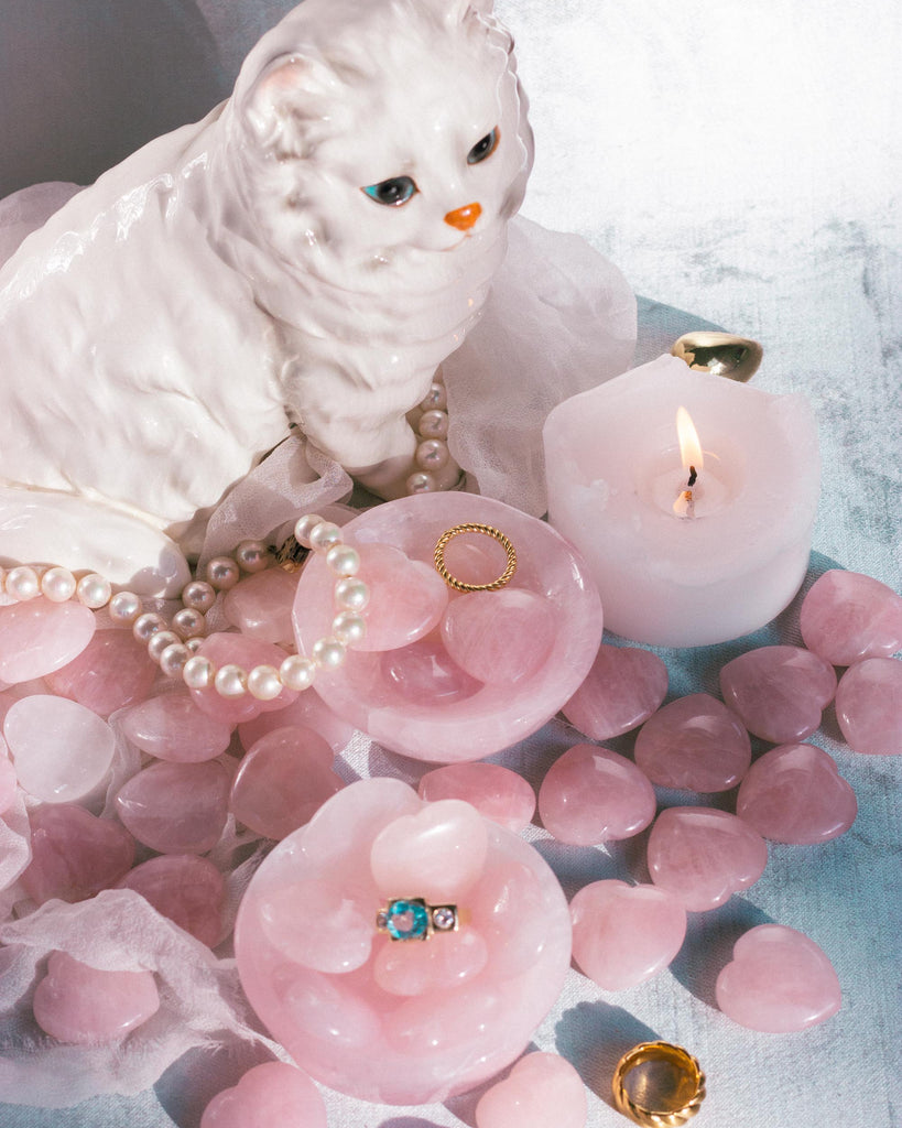 A rose quartz crystal bowl holding smooth, hand-carved natural Rose Quartz crystal worry stone hearts, set on textured blue velvet with scattered gold jewelry and a cat.