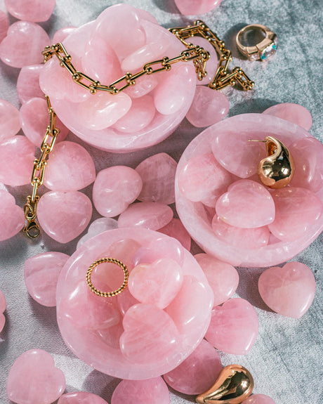 A rose quartz crystal bowl containing natural polished Rose Quartz Crystal Hearts surrounded by similar pinkish crystal hearts and gold jewelry on a textured velvet blue background.