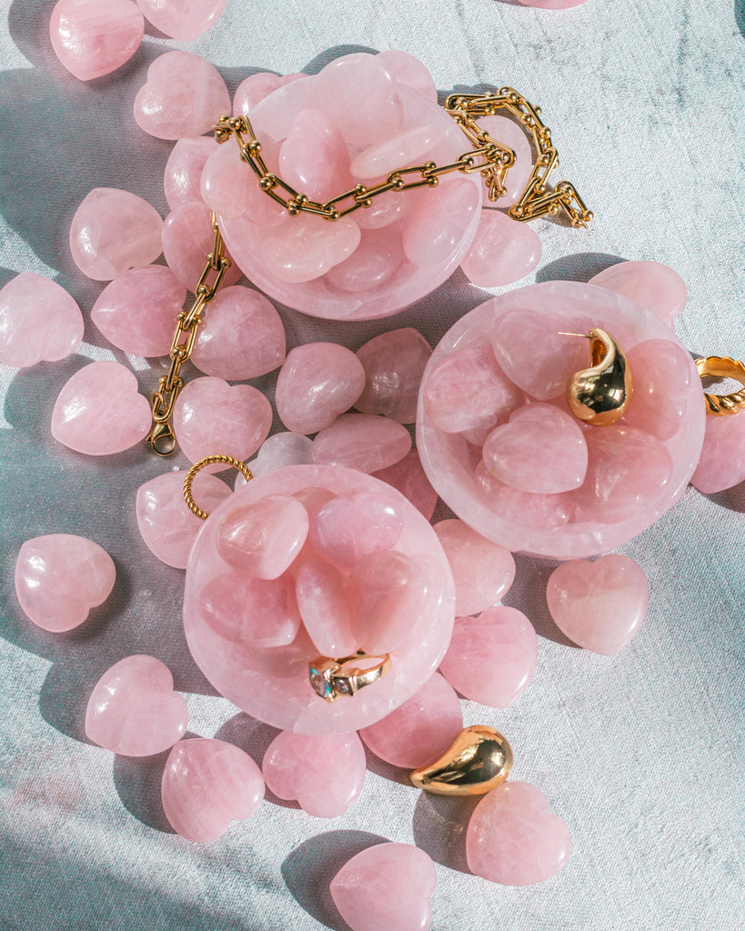 Hand-carved 100% natural Rose Quartz crystal hearts displayed in rose quartz bowls on textured navy velvet, surrounded by gold jewelry accents