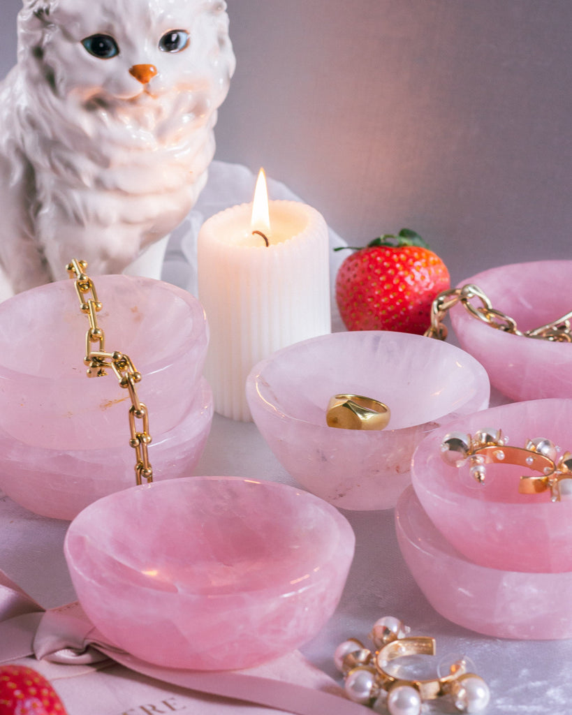 Pink jewelry rose quartz crystal bowls with gold jewelry, strawberries, and a lit candle on a light background.