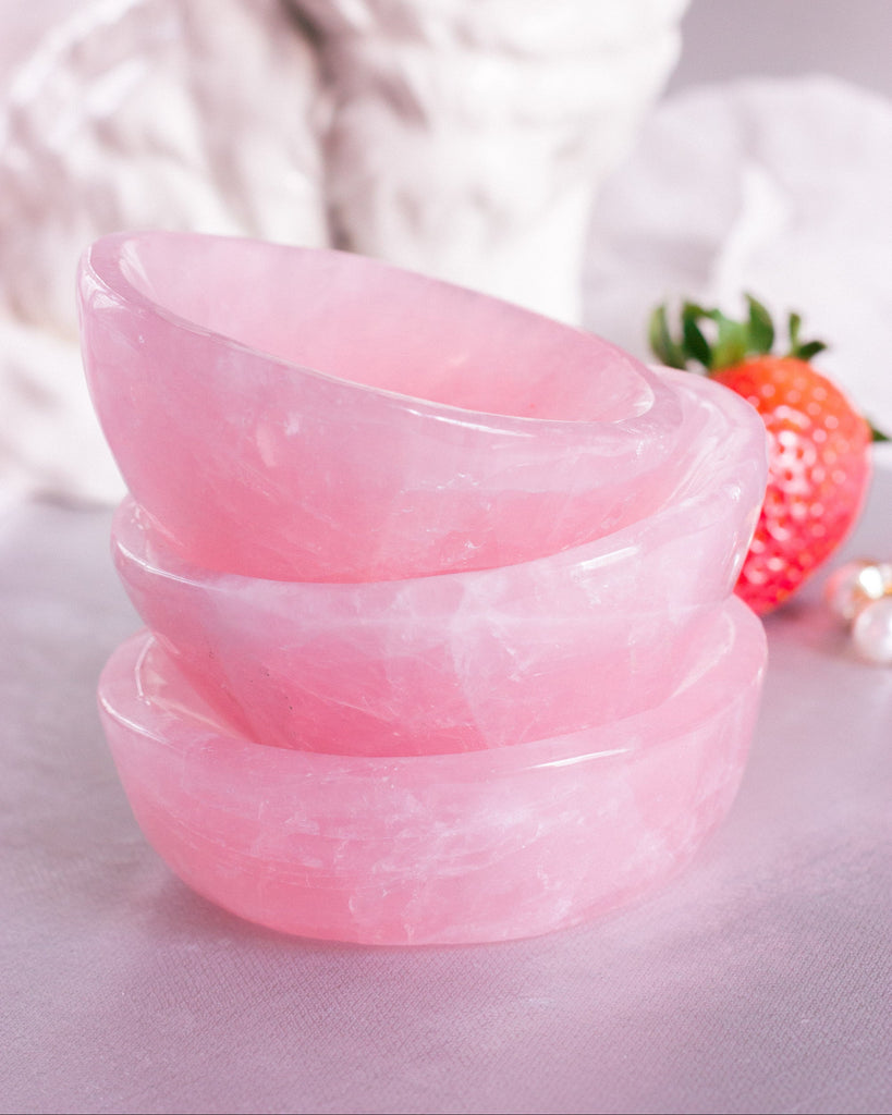 Stacked Hand-carved Rose Quartz crystal dishes with soft pink hues, displayed on a blue velvet background with crystal energy stones nearby  and strawberries.