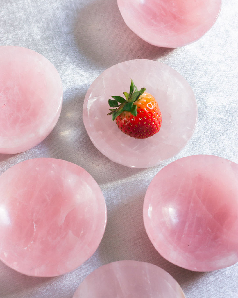 Natural hand-carved Rose Quartz crystal dish with smooth polished surface, styled on blue velvet with heart chakra crystals nearby and a strawberry.